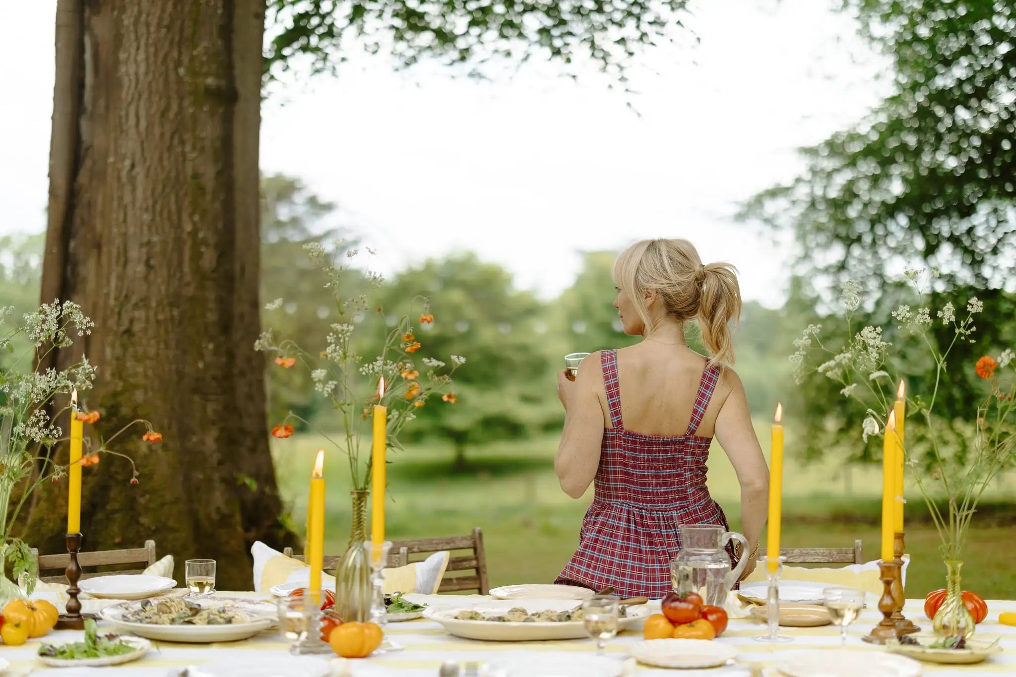 Clodagh McKenna leaning on an outdoor table with candles and flowers, looking at a scenic view.