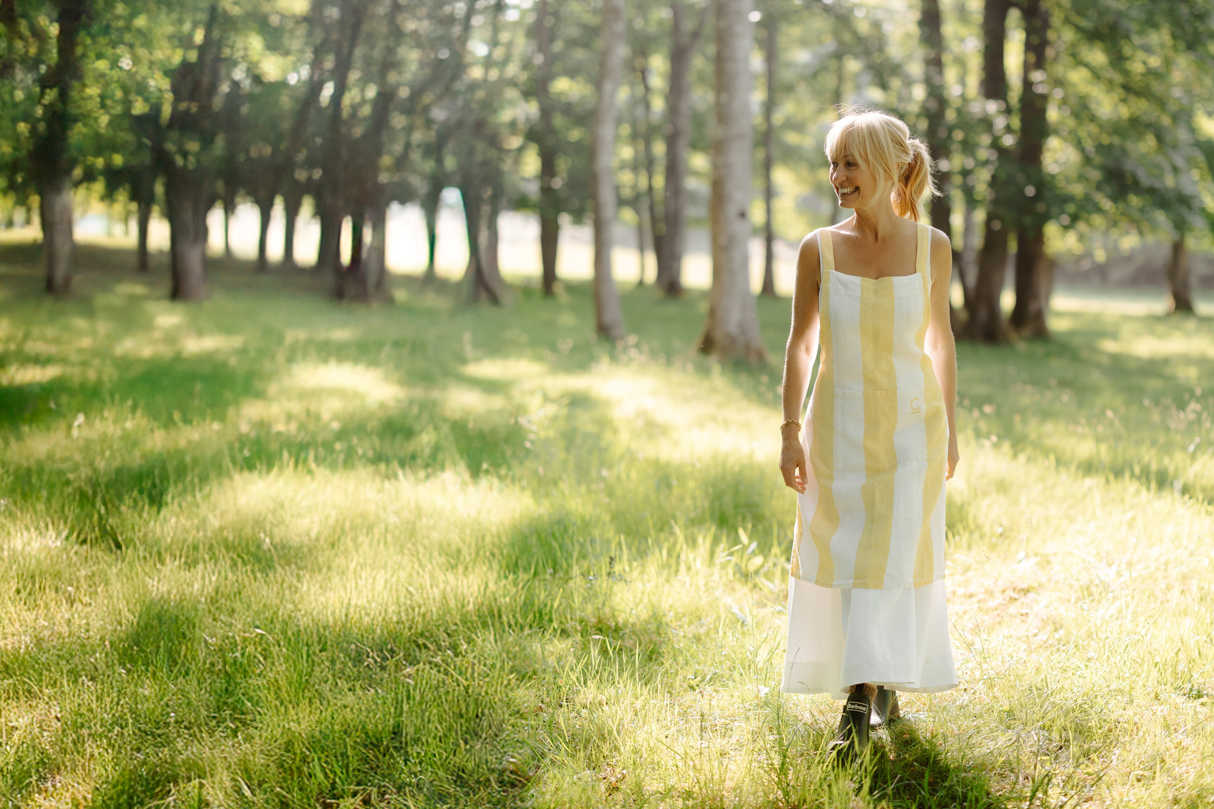 Clodagh McKenna in a yellow and white striped apron standing in a sunlit meadow.