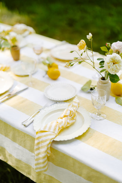 Outdoor table setting with yellow and white striped tablecloth, plates, and flowers.