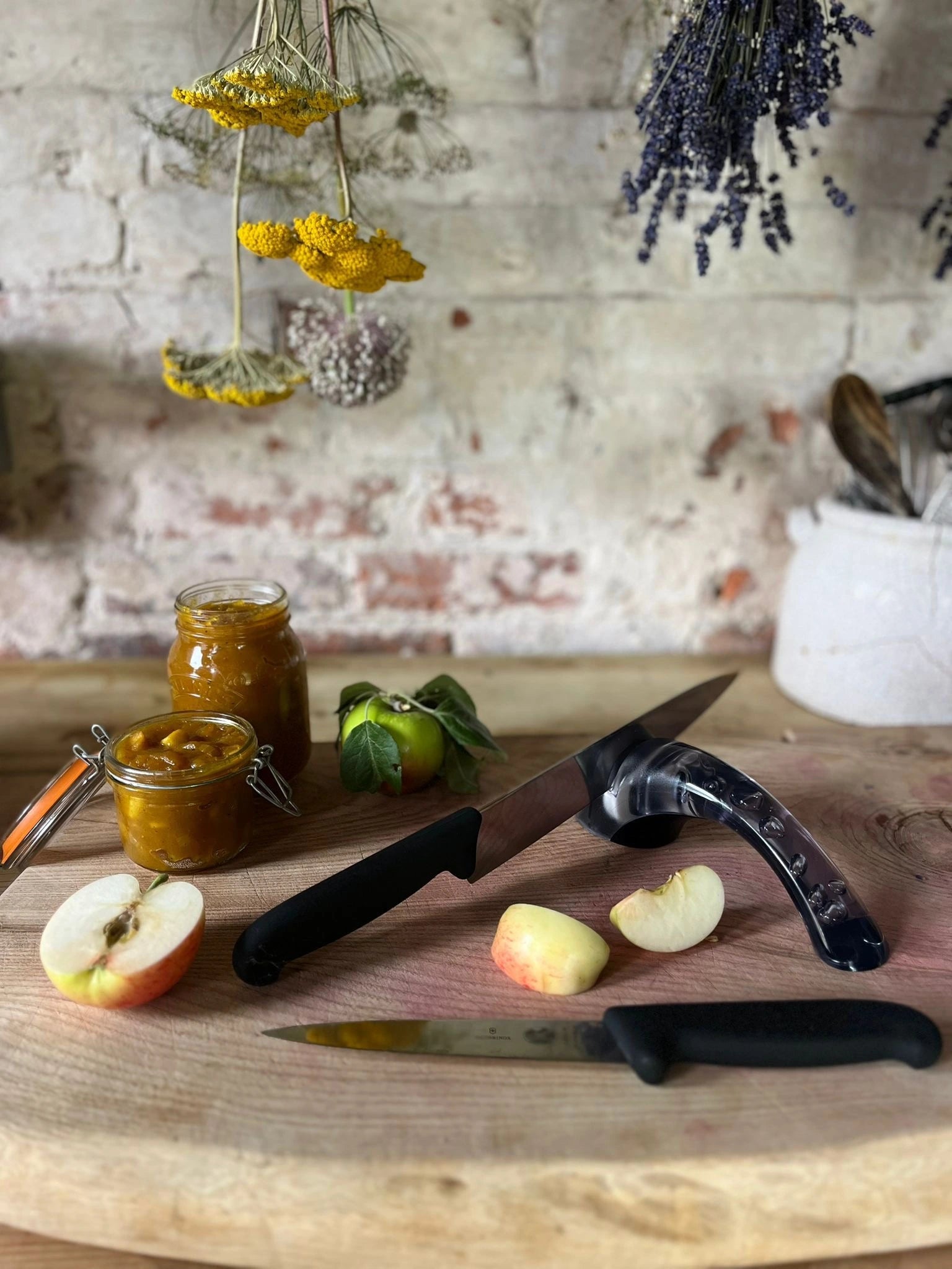 Wooden cutting board with jars of preserves, apples, and knives and a knife sharpener against a rustic brick wall.